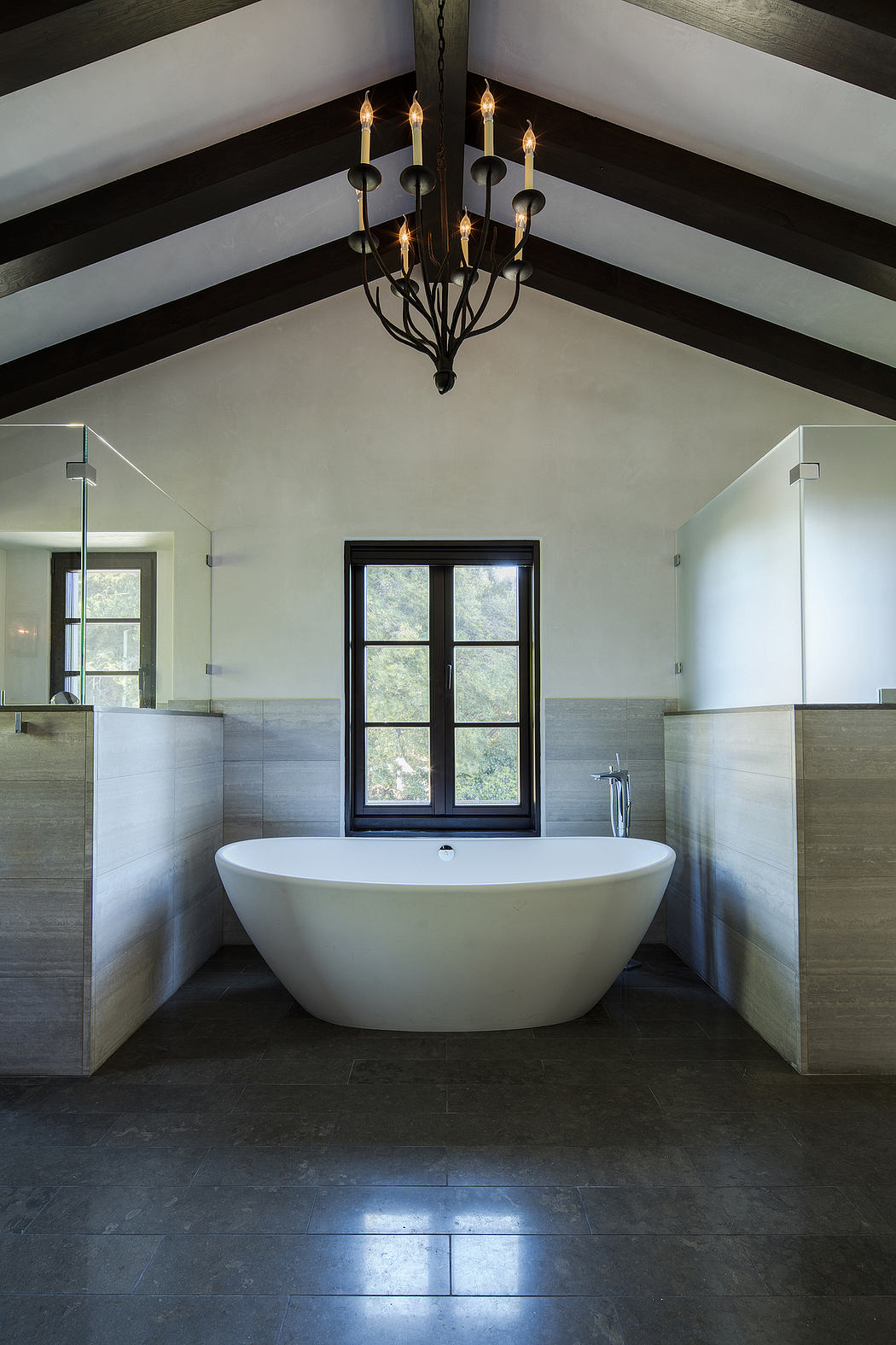 A modern, minimalist bathroom with a freestanding tub, black-framed window, and a chandelier overhead.