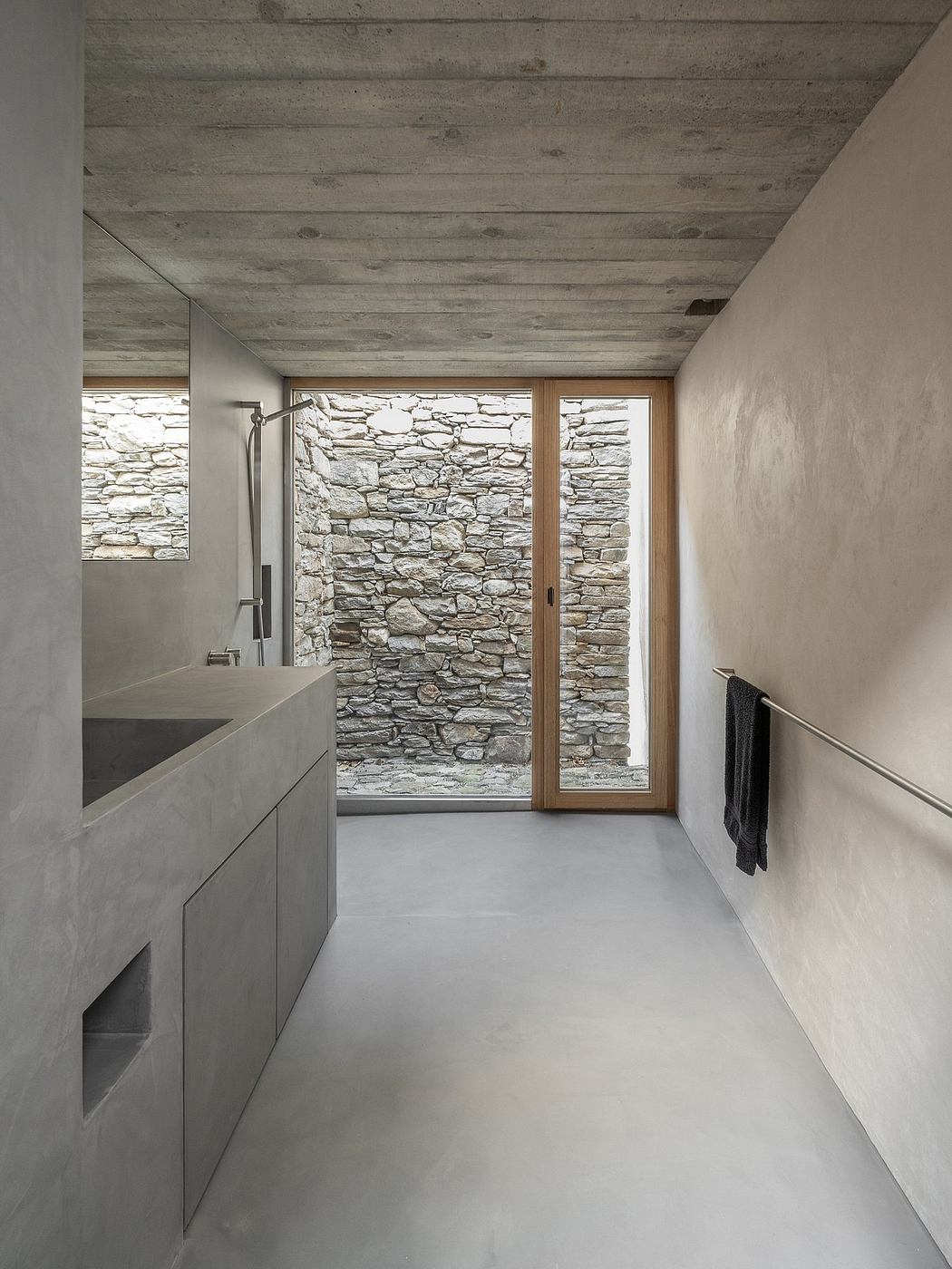 A modern bathroom with concrete floors, stone walls, and a wooden door leading to the exterior.