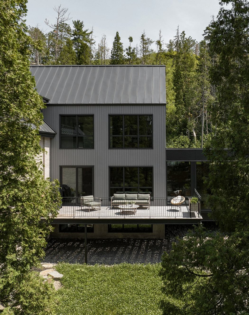 A modern, two-story house with a black metal roof and expansive outdoor deck surrounded by lush greenery.