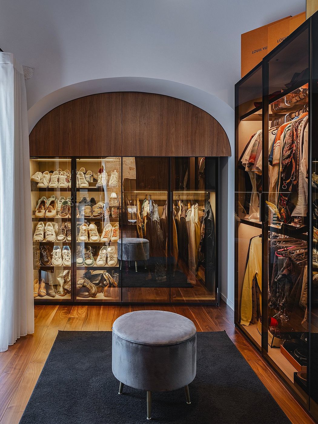 A spacious walk-in closet with wooden arched wall, glass shelves, and a grey ottoman.