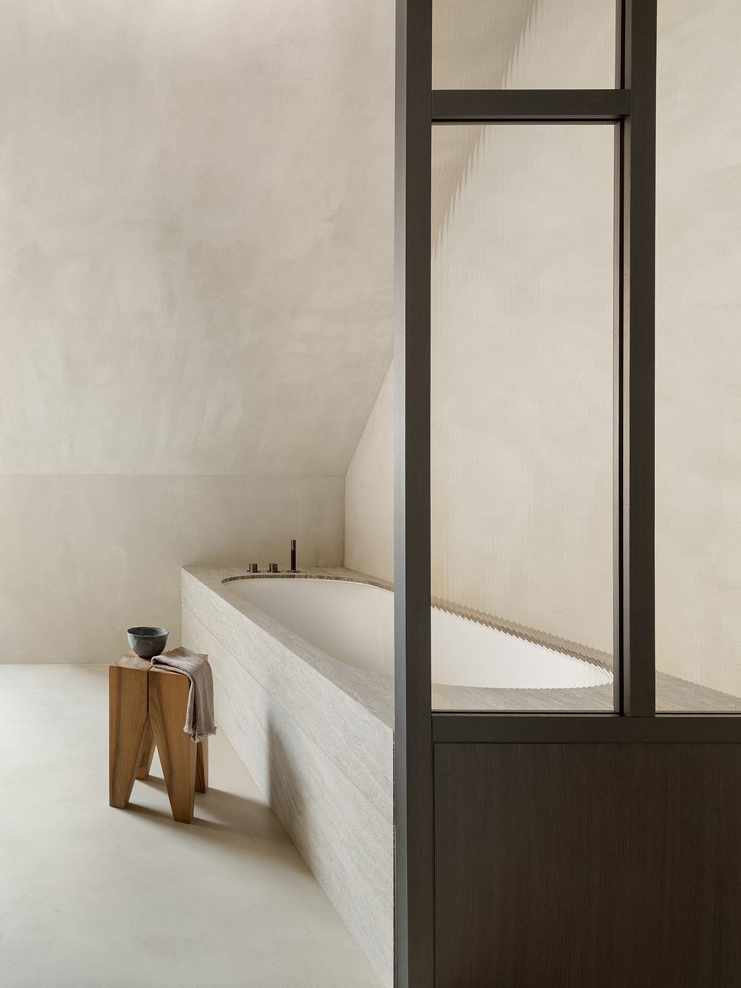 A minimalist bathroom with a freestanding bathtub and a wooden stool, framed by a steel and glass wall.