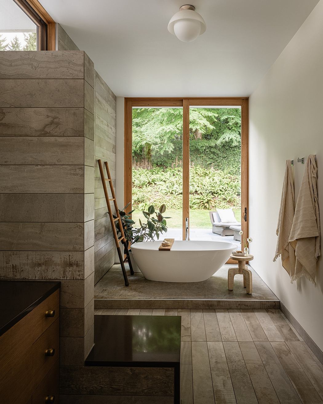 Serene bathroom with freestanding tub, wood-paneled walls, and large windows overlooking lush greenery.