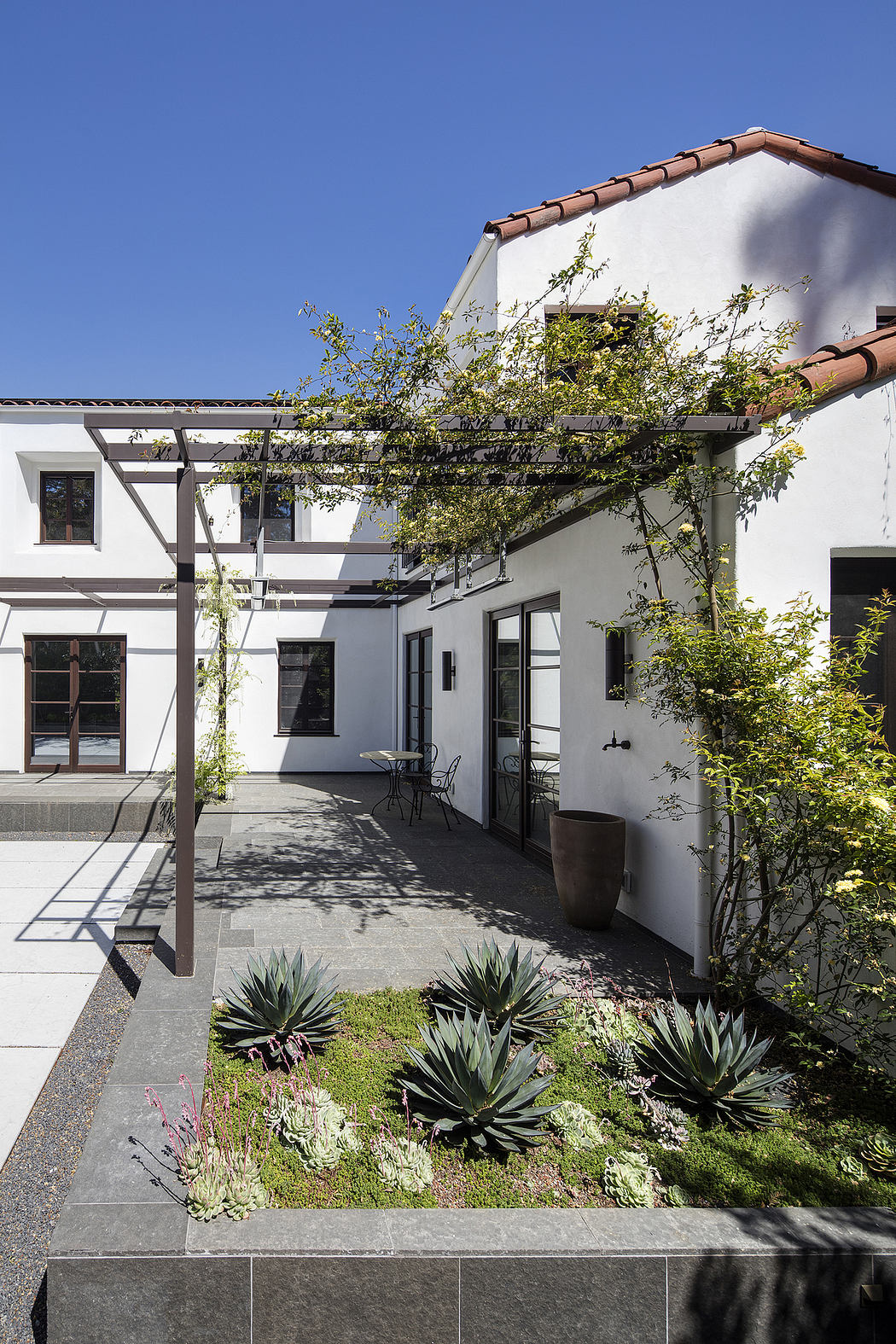 A spacious courtyard with a modern, white-washed exterior, lush plants, and a pergola.