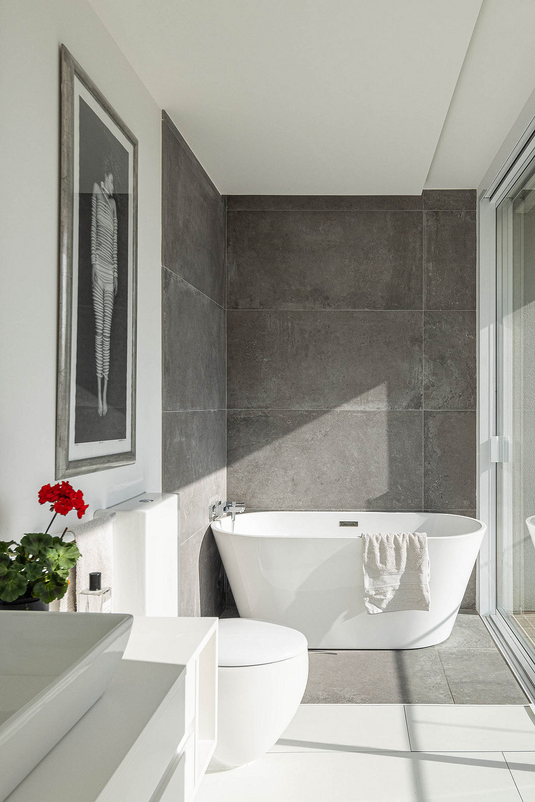 A modern bathroom with a white freestanding tub, gray tile walls, and a framed artwork.