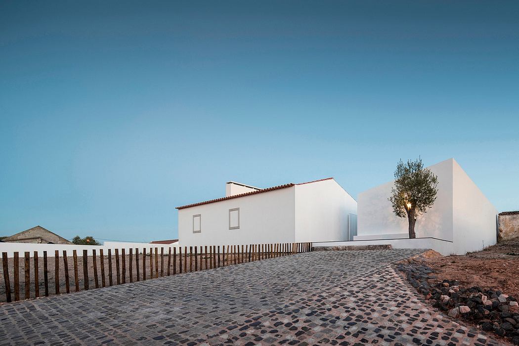 A modern white building with clean lines and a wooden fence leading to a cobblestone path and a lone tree.