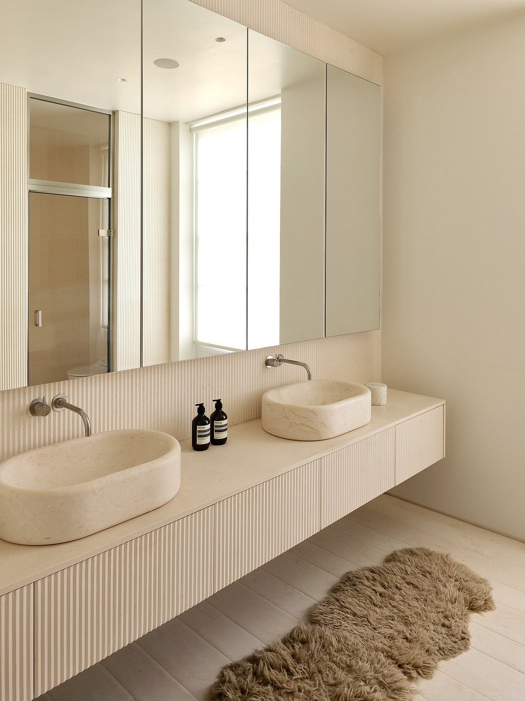 Minimalist bathroom with beige tile, stone sinks, and a large wall mirror. Cozy sheepskin rug completes the serene look.