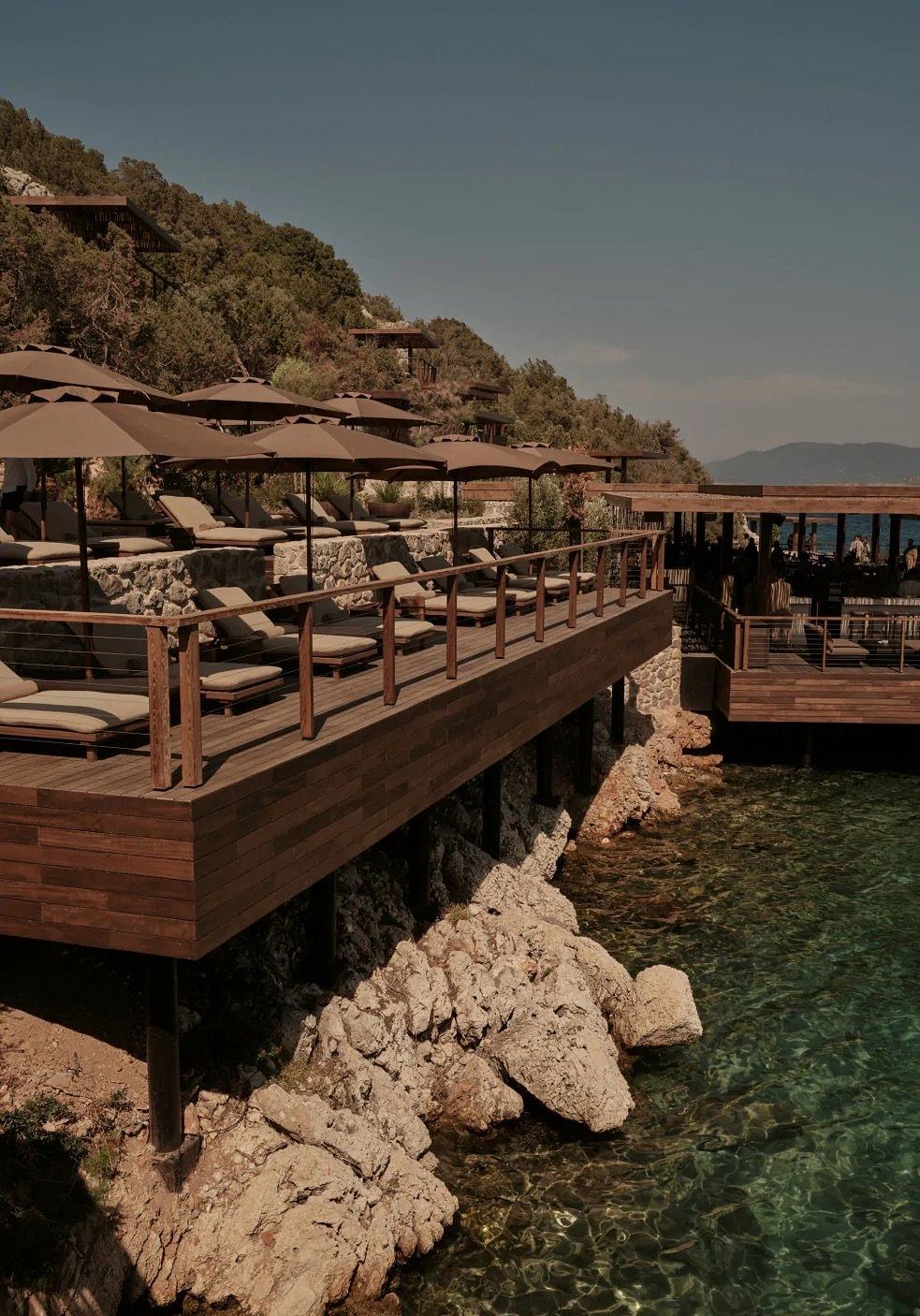A wooden deck with umbrellas and lounge chairs overlooking a rocky coastline and turquoise waters.