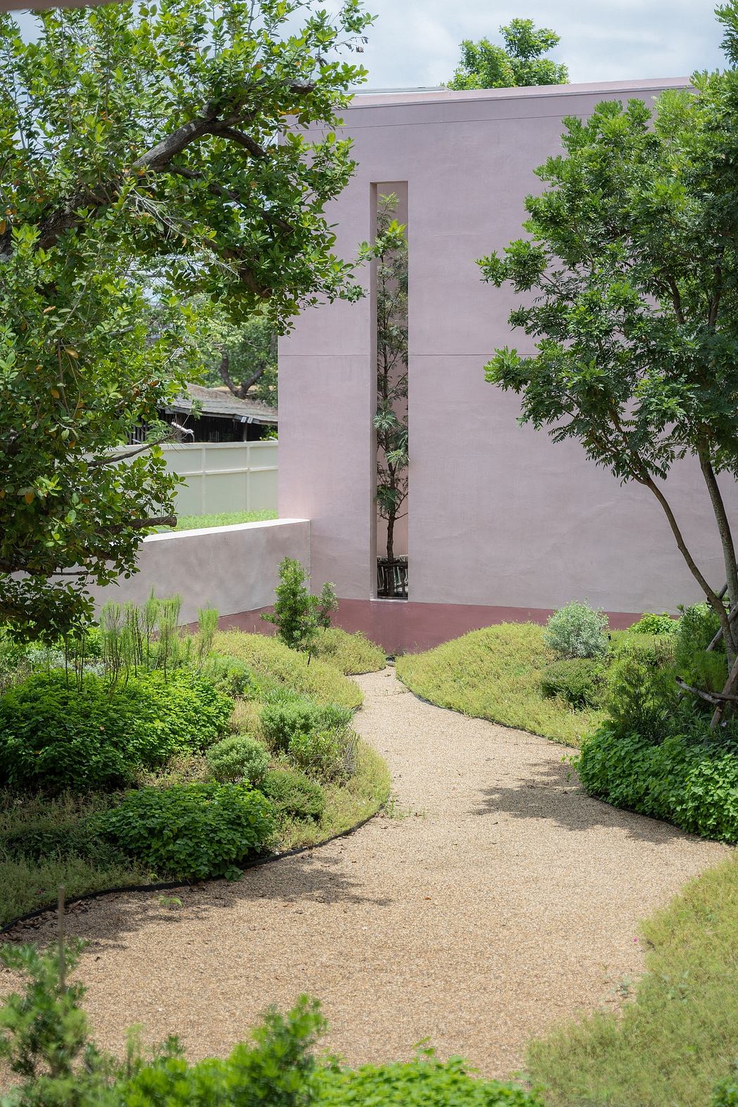 A serene outdoor garden path leads to a modern, minimalist building with a pink-hued facade.
