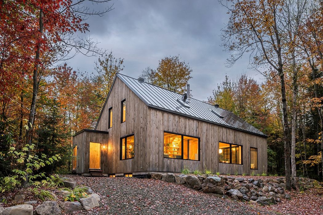 Modern wooden cabin with a gabled roof, large windows, and a stone foundation nestled in a fall forest.