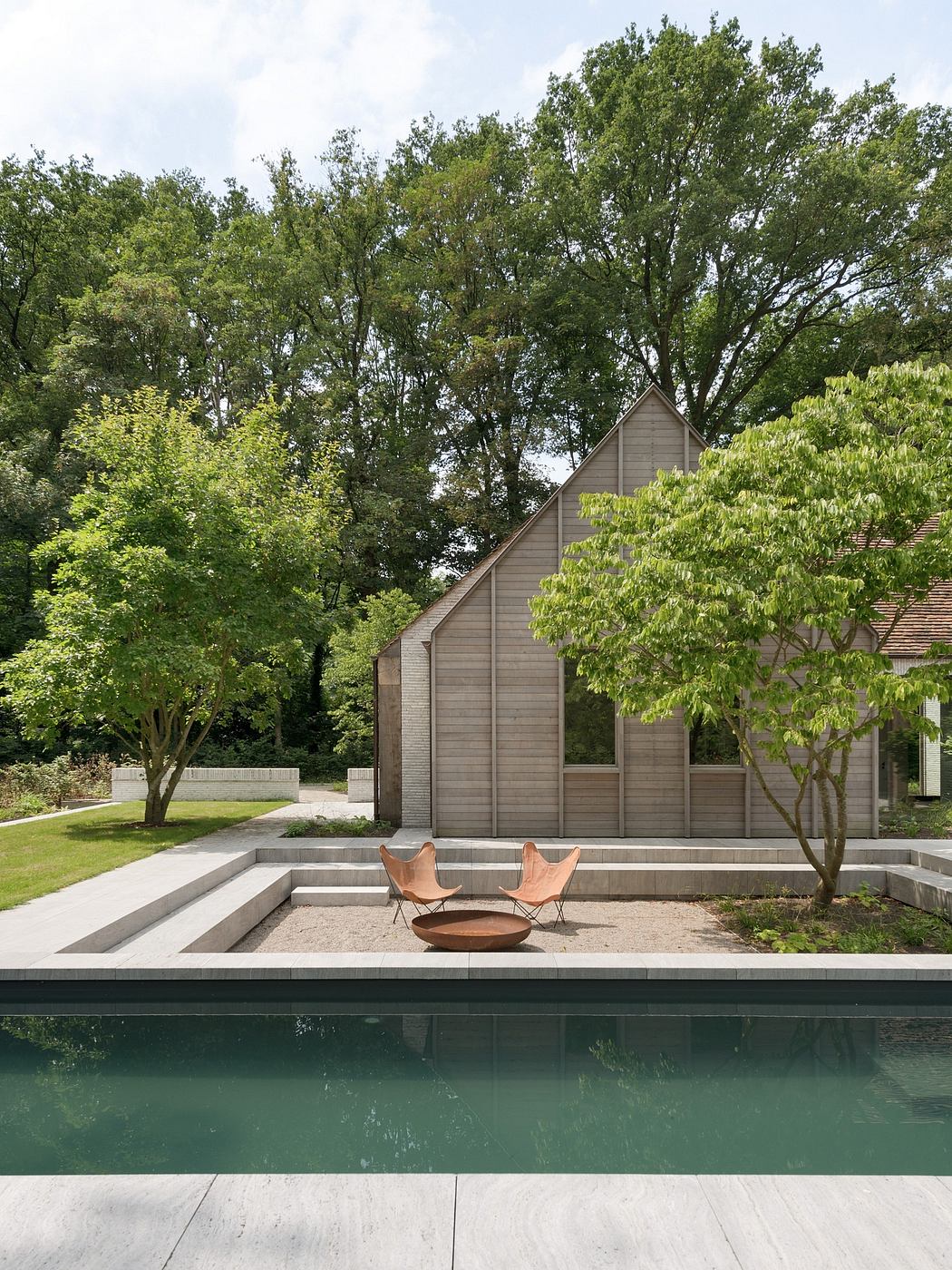 A modern wooden structure with a pool and two rustic chairs, surrounded by lush greenery.