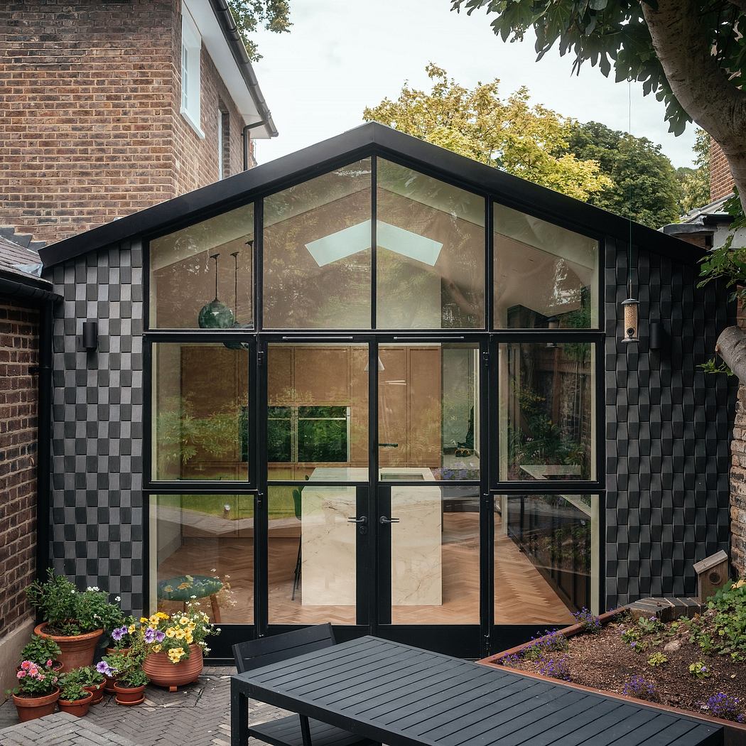 A modern, glass-enclosed structure with black metal framing, set against a brick backdrop and surrounded by lush foliage.