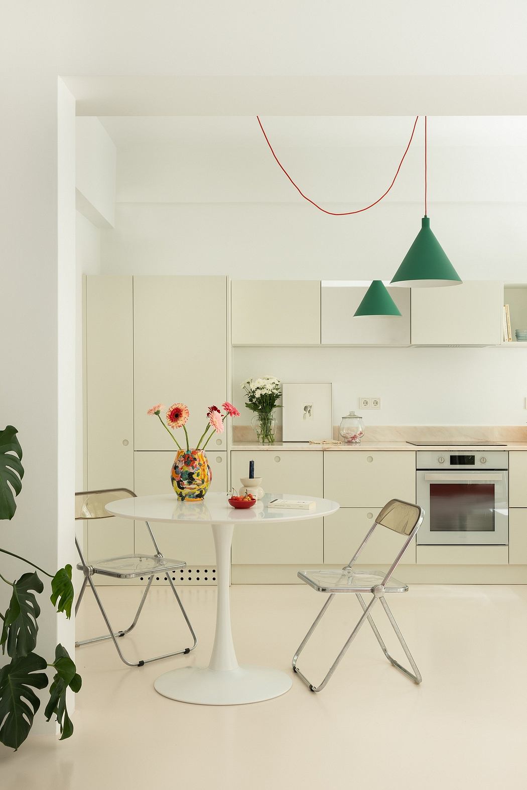 A minimalist kitchen with white cabinets, green pendant lights, a round table, and chrome chairs.