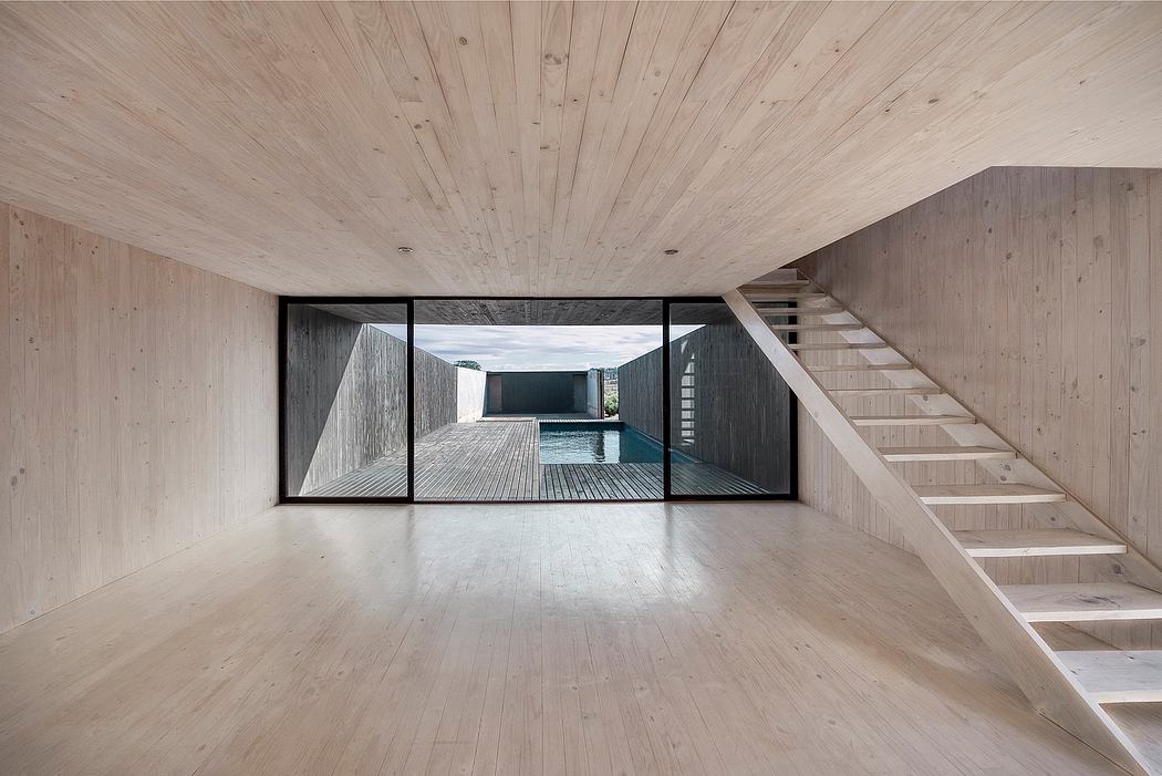Minimalist open-plan interior with wooden paneled walls, floor, and stairs. Expansive glass doors lead to a deck.