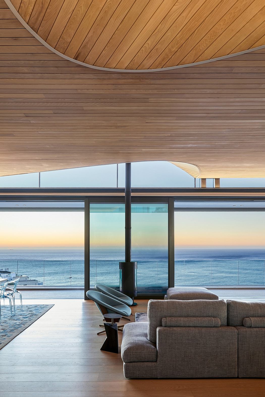Modern beach house with curved wood ceiling, floor-to-ceiling windows, and plush seating.