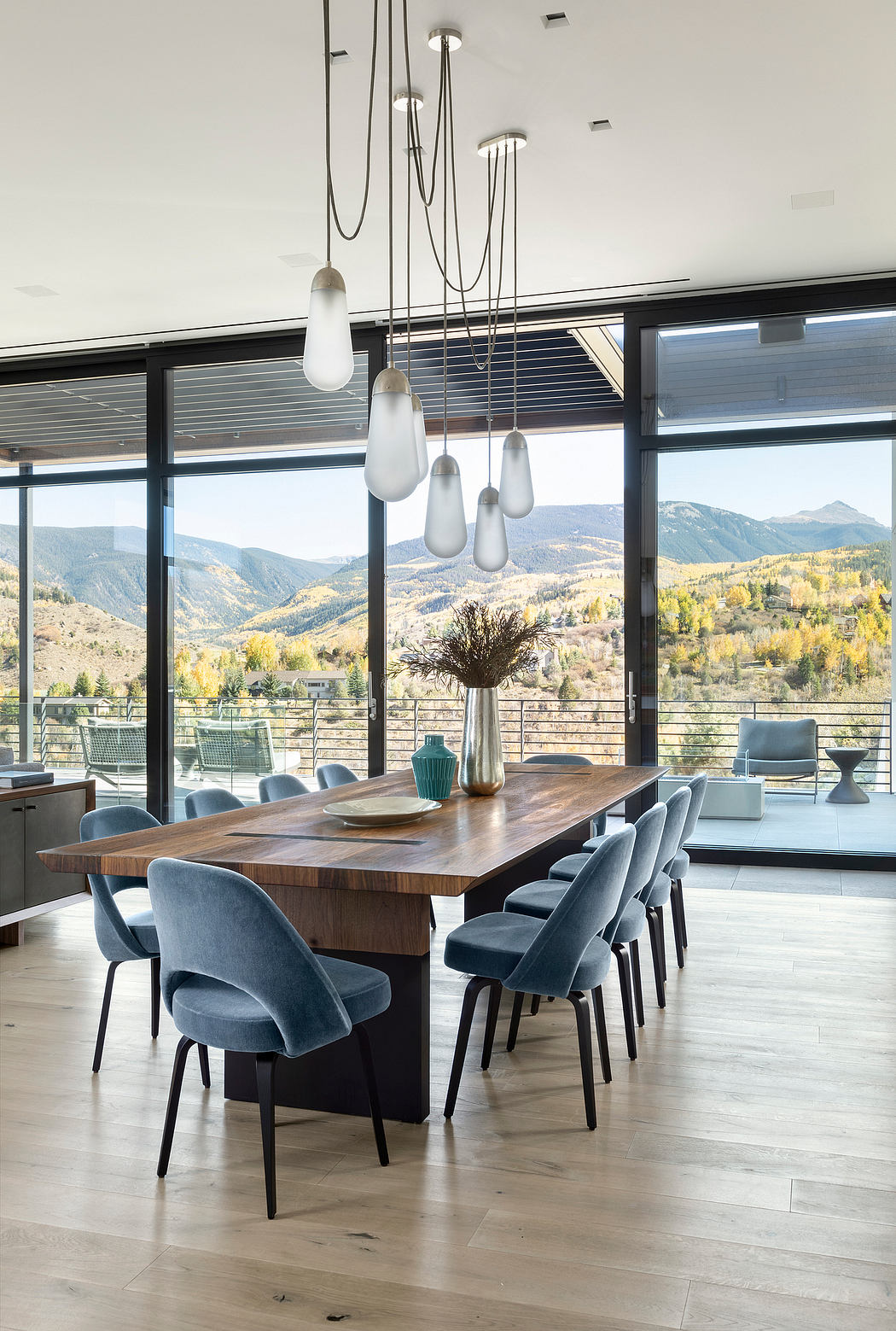 Contemporary dining room with wooden table, grey velvet chairs, and pendant lights; expansive mountain view through large windows.