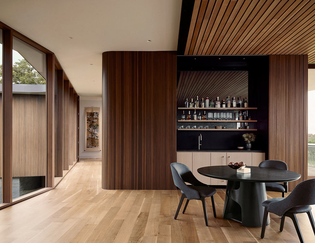 A modern open-plan living space with warm wood paneling, a bar, and a circular dining table.