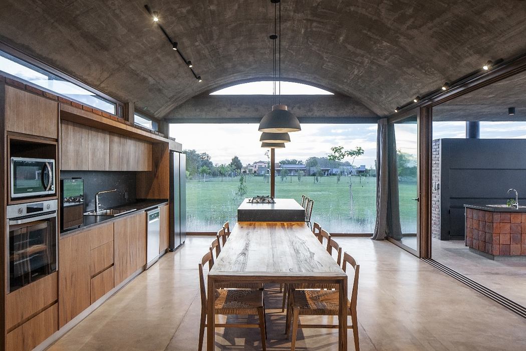 Modern open-plan kitchen and dining area with wooden furniture, concrete ceiling, and large windows offering scenic views.