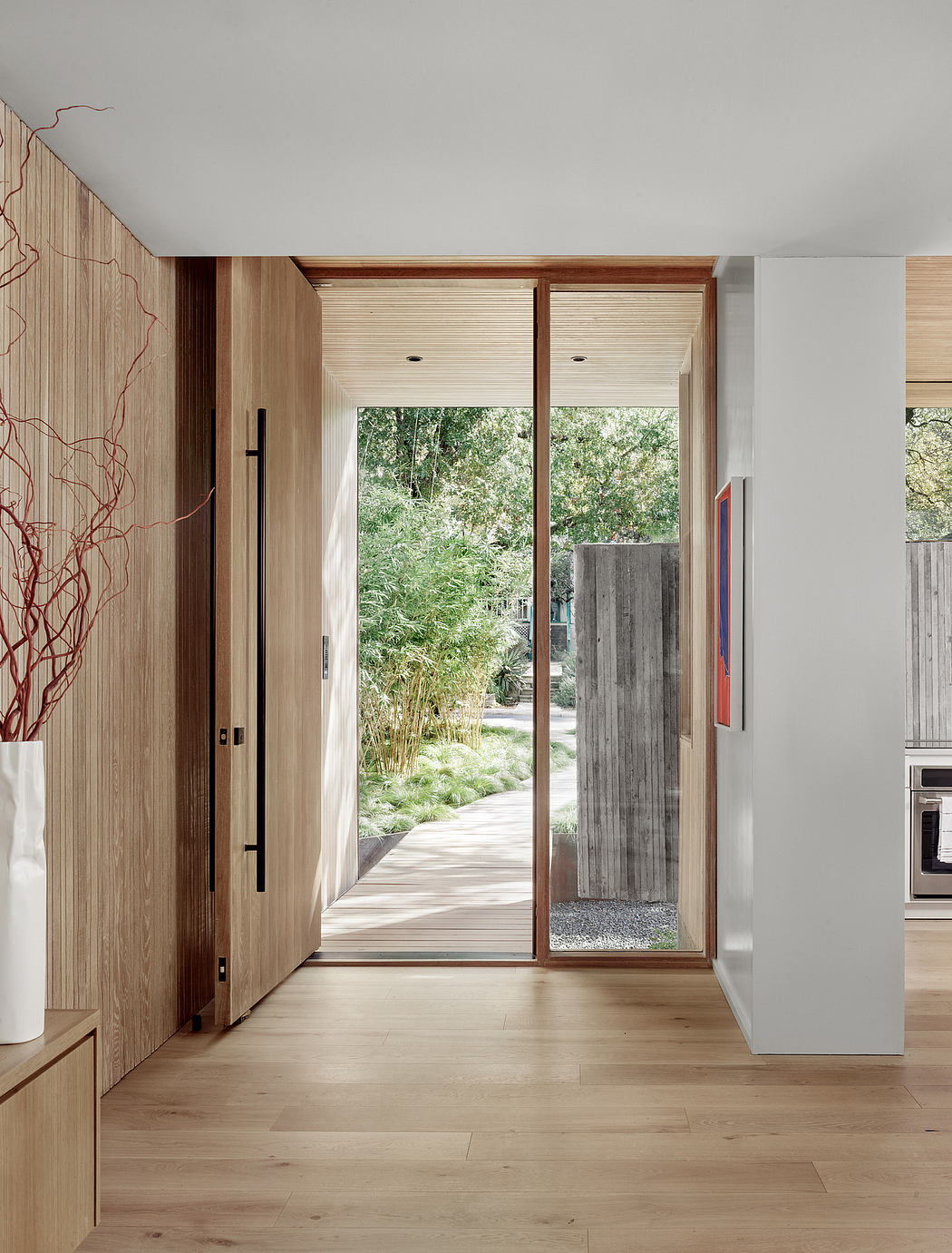 The image features a modern, minimalist entryway with a large sliding glass door, wooden accents, and a lush outdoor view.