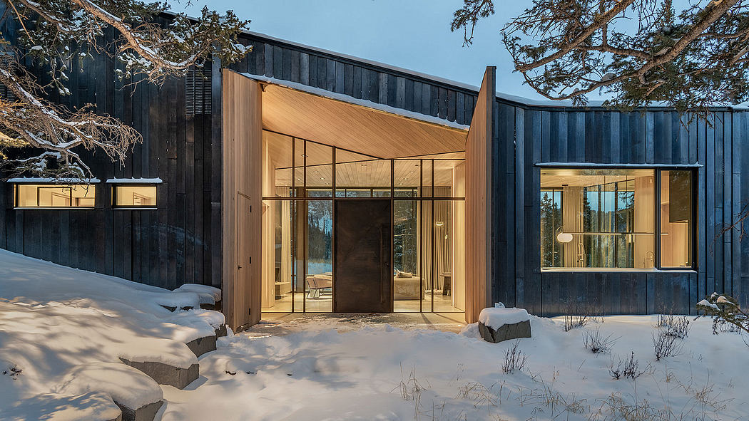A modernist winter cabin with wood and glass facade, stone walkway, and snowy surroundings.