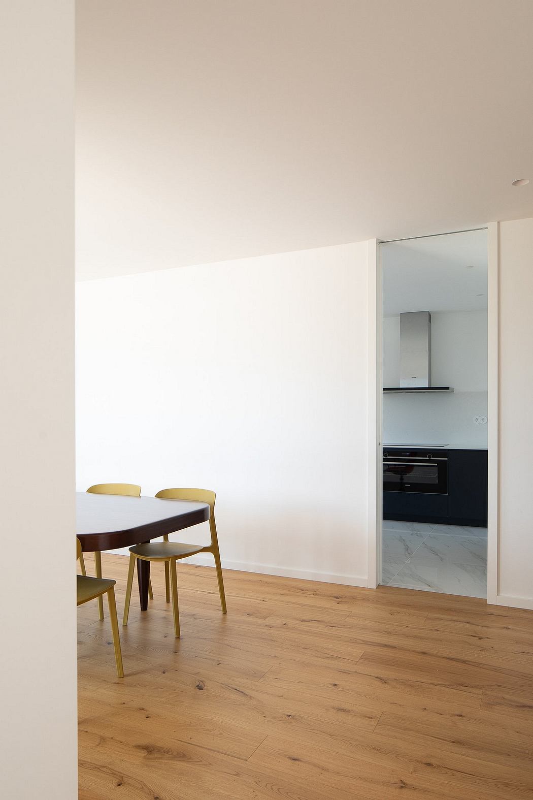 Modern, minimalist dining area with wooden flooring, white walls, and built-in kitchen appliances.