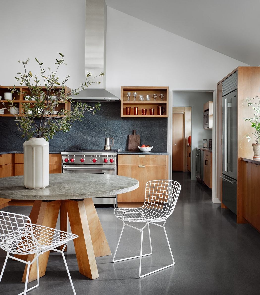 A modern kitchen with wooden cabinets, a sleek range, and stylish wire chairs around a concrete table.