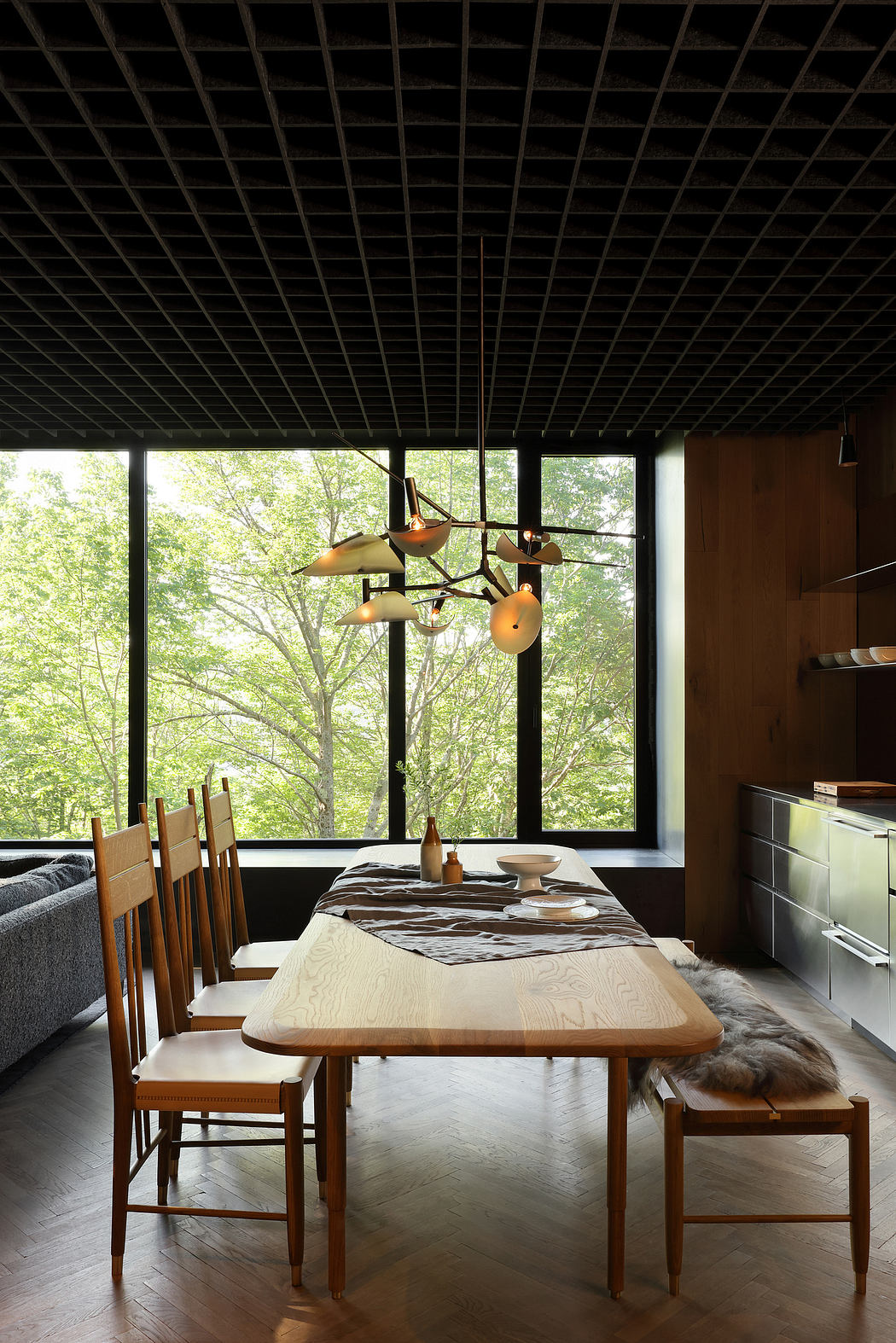 A modern, nature-inspired dining room with a large wooden table, mid-century lighting, and floor-to-ceiling windows.
