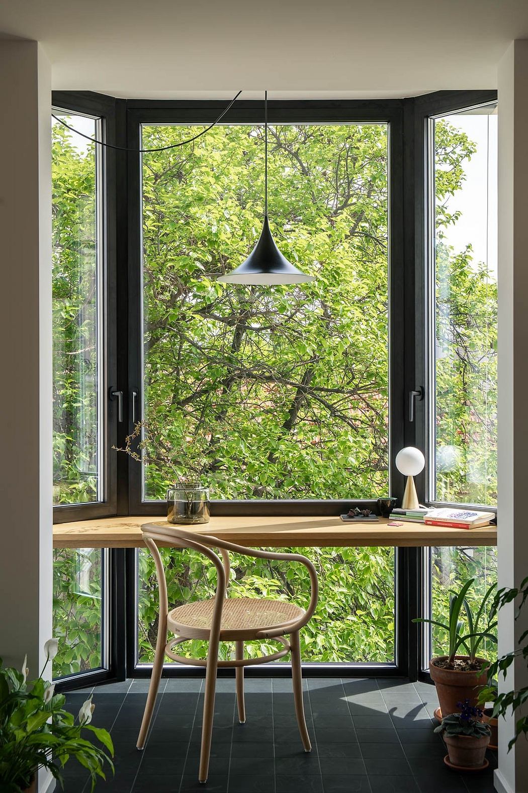 A cozy window nook with a wooden desk, minimalist pendant light, and lush greenery outside.