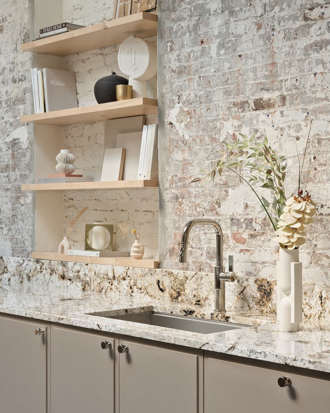 A modern kitchen with white shelves, a granite countertop, and a rustic brick wall.