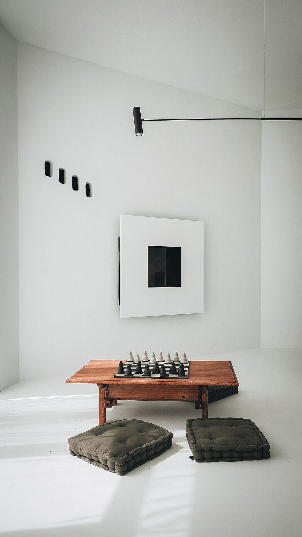 A minimalist room with a wooden table, chess set, and padded floor cushions. Geometric lighting and artwork.
