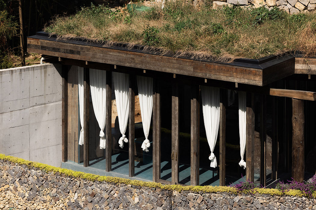 A rustic wooden structure with glass windows and billowing white curtains, surrounded by a grassy landscape.