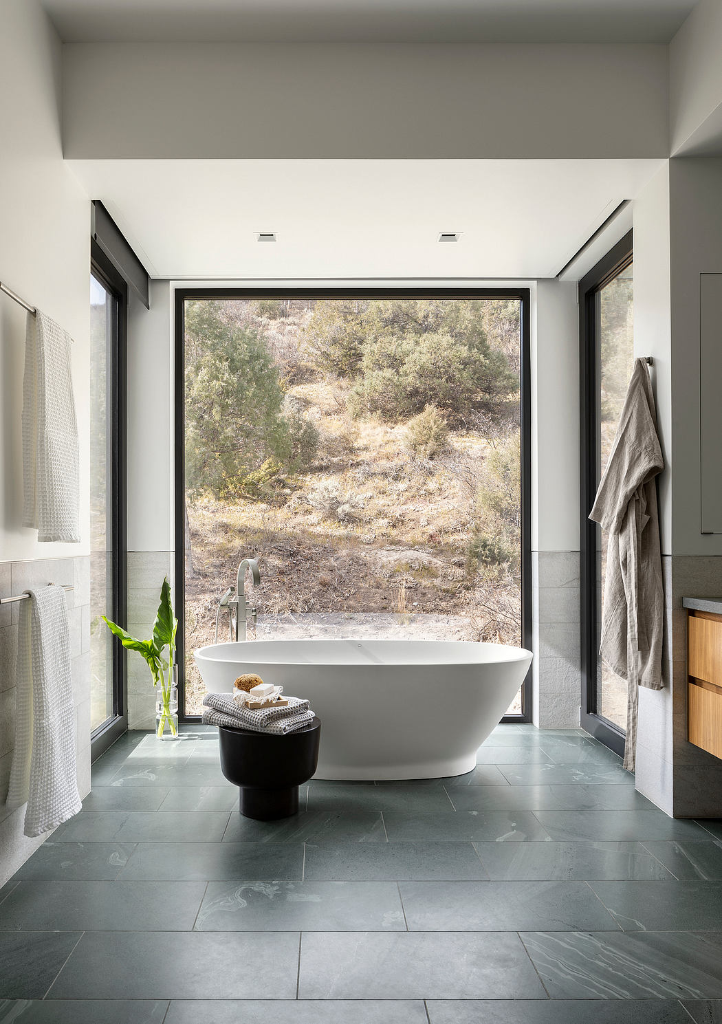 A modern bathroom with a freestanding tub, large windows overlooking a forested landscape.