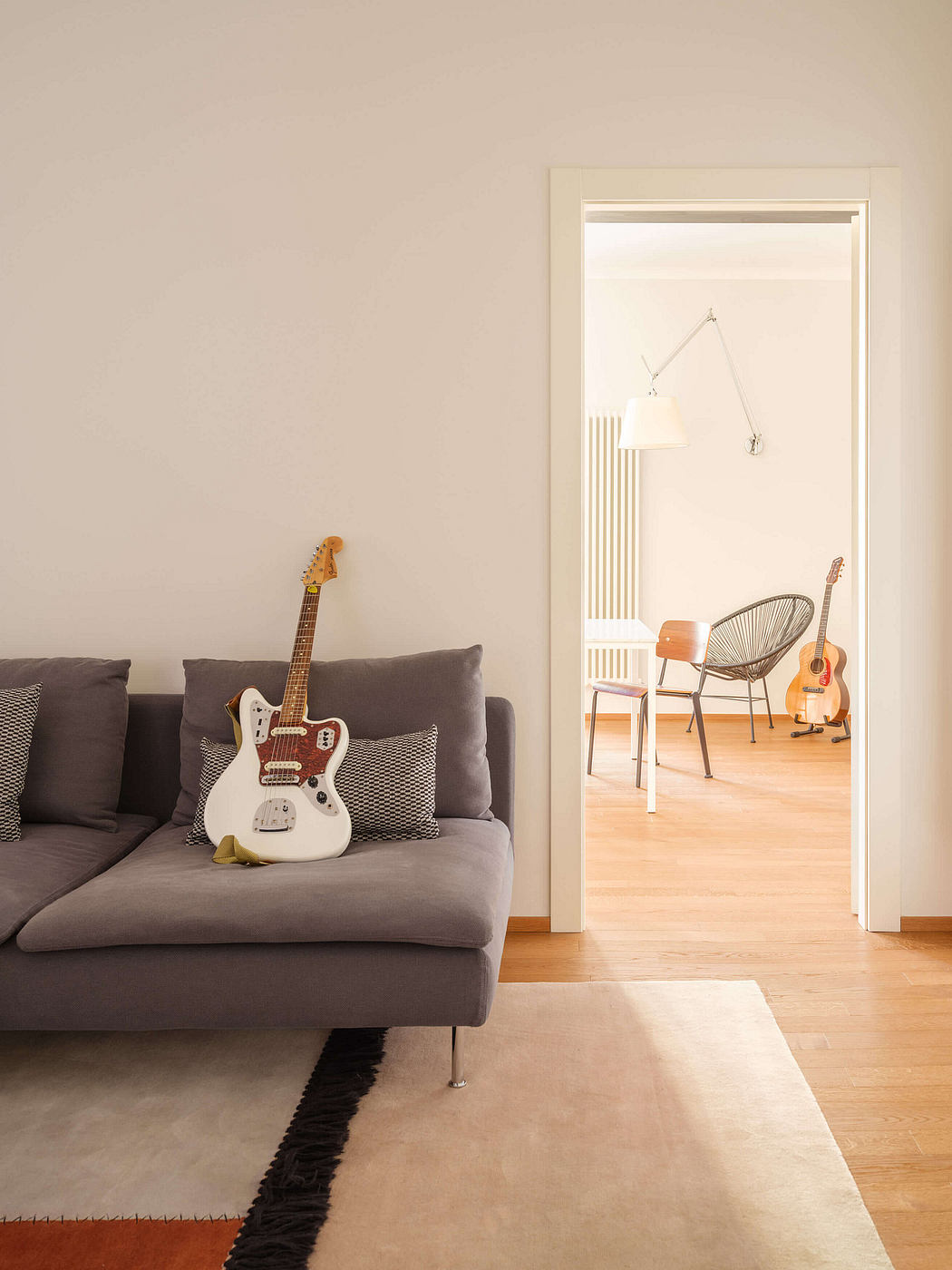 Minimalistic living room with grey sofa, white guitar, and open doorway revealing a chair.