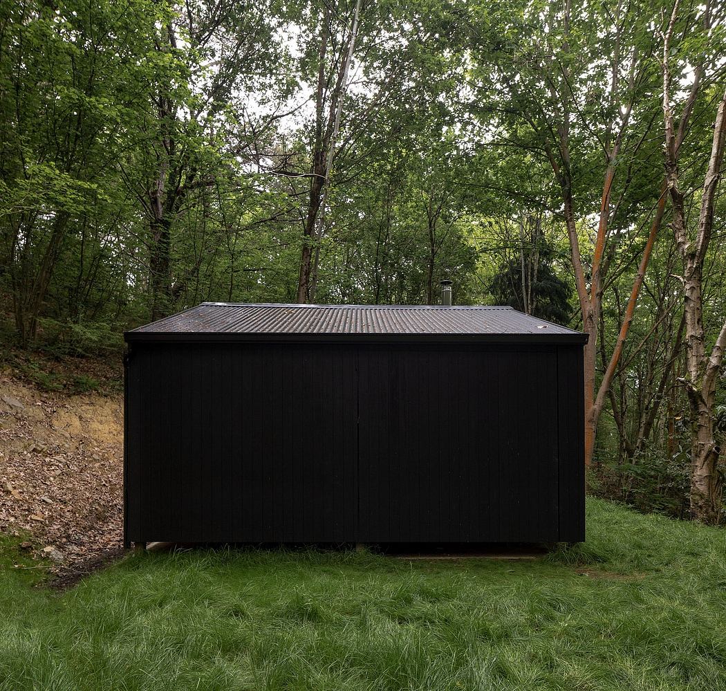 A black, minimalist structure nestled in a lush, green forest, blending seamlessly with the natural surroundings.