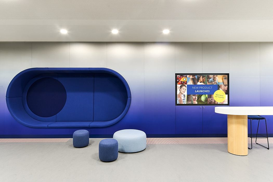 Modern office lounge with blue seating, video display, and minimalist table and stools.