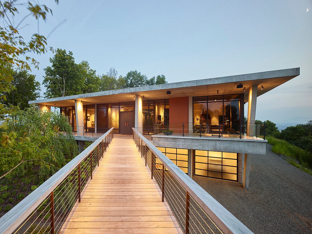 Modern wooden bridge leads to a large, open-concept house with glass walls and a slanted roof.
