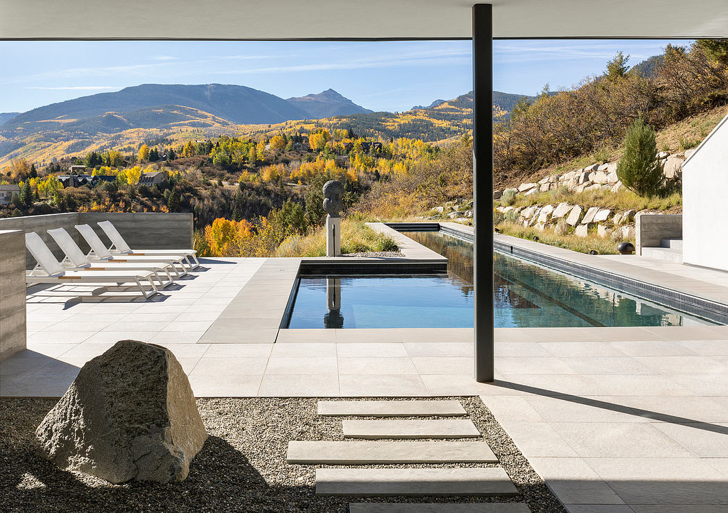 The image shows a modern, minimalist outdoor space with a sleek infinity pool, lounge chairs, and a stunning mountain landscape backdrop.
