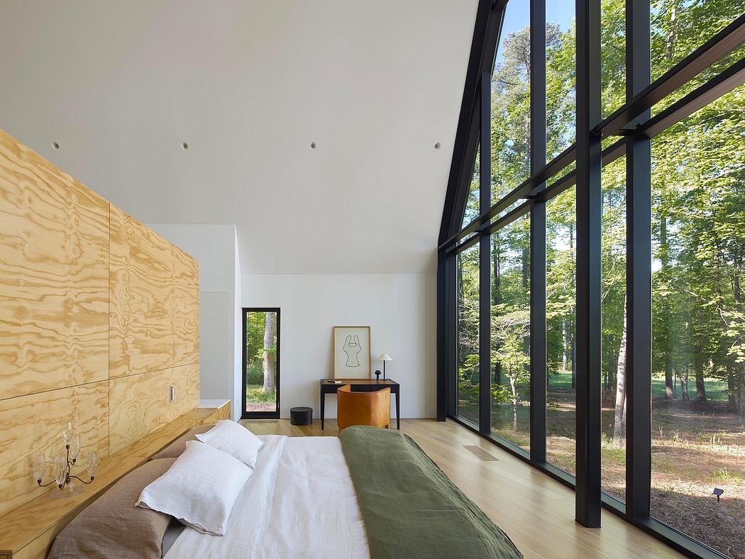 A spacious modern bedroom with large windows overlooking a forested landscape. Plywood paneling, minimalist furniture.