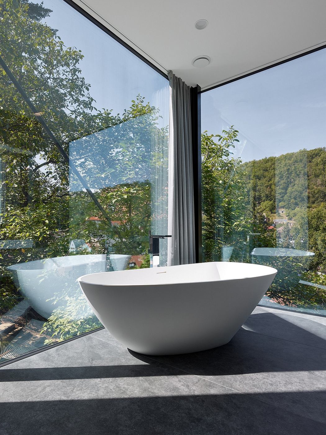 A modern freestanding bathtub overlooking a lush, forested outdoor scene through floor-to-ceiling glass walls.