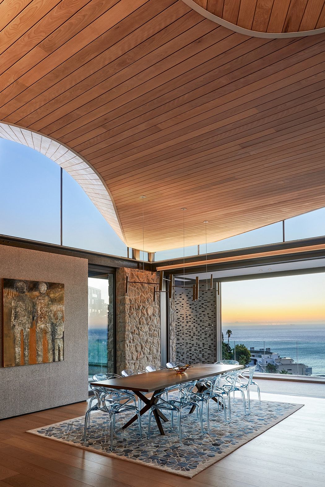 Modern, open-concept dining area with wooden ceiling, stone wall, and panoramic ocean view.