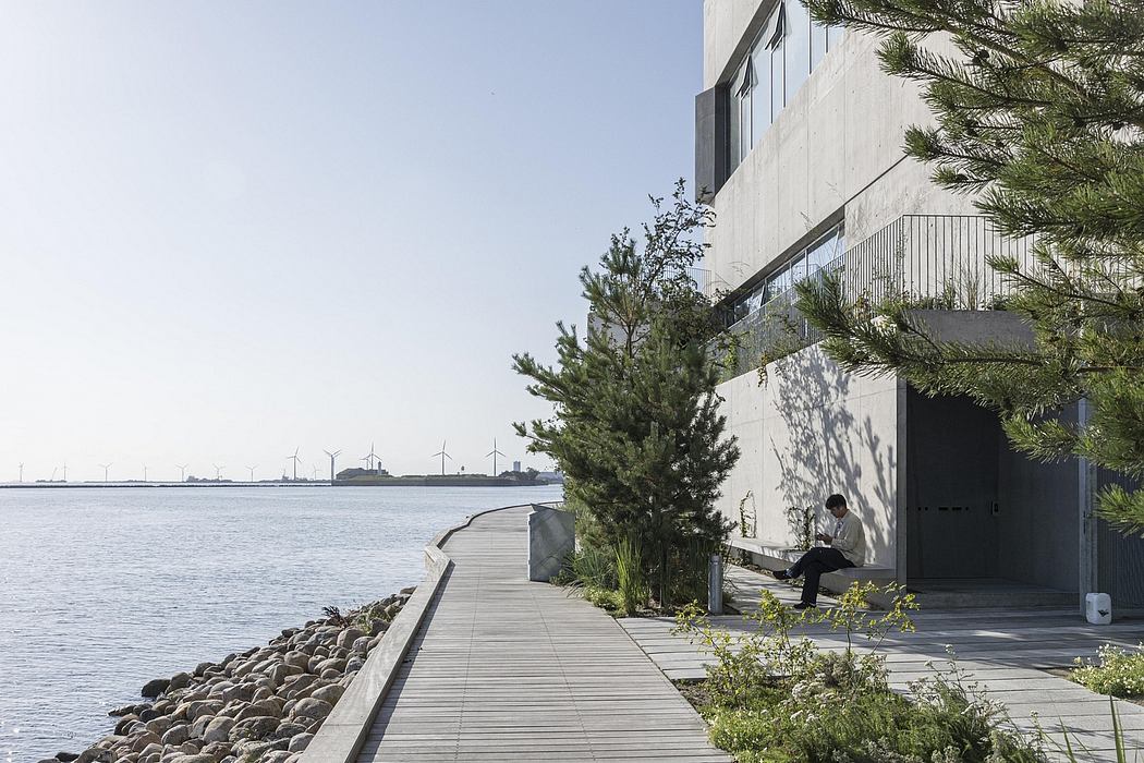 A sleek, modern building with a wooden boardwalk leading to the waterfront, featuring wind turbines on the horizon.
