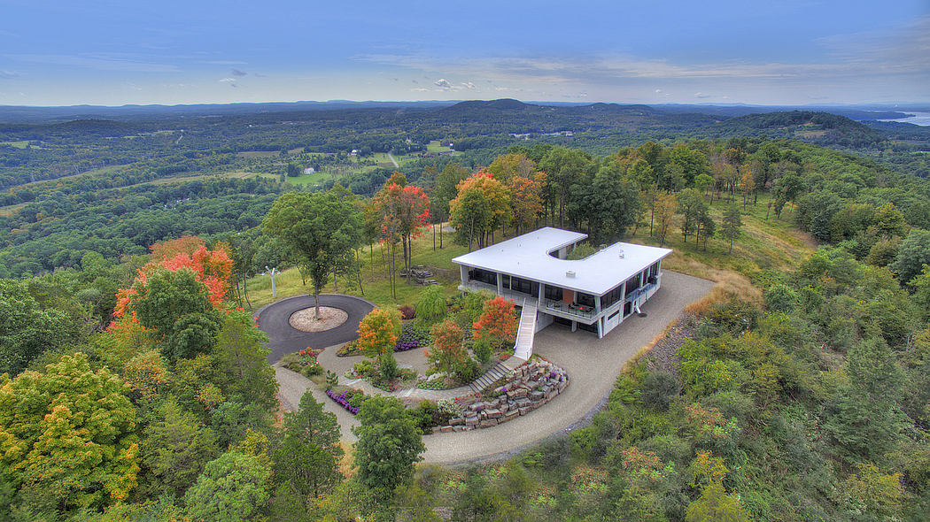 A modern hilltop cabin with a wraparound porch overlooking a lush, colorful landscape.