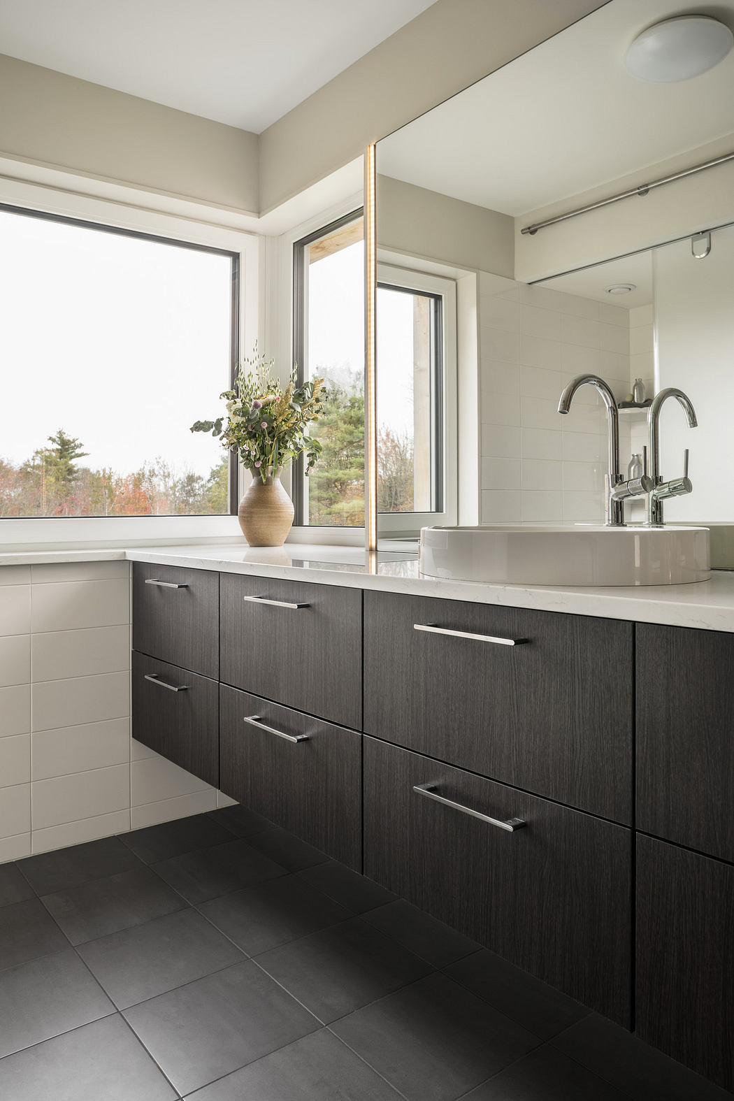 A modern bathroom with dark wood cabinets, a white countertop, and large windows overlooking nature.