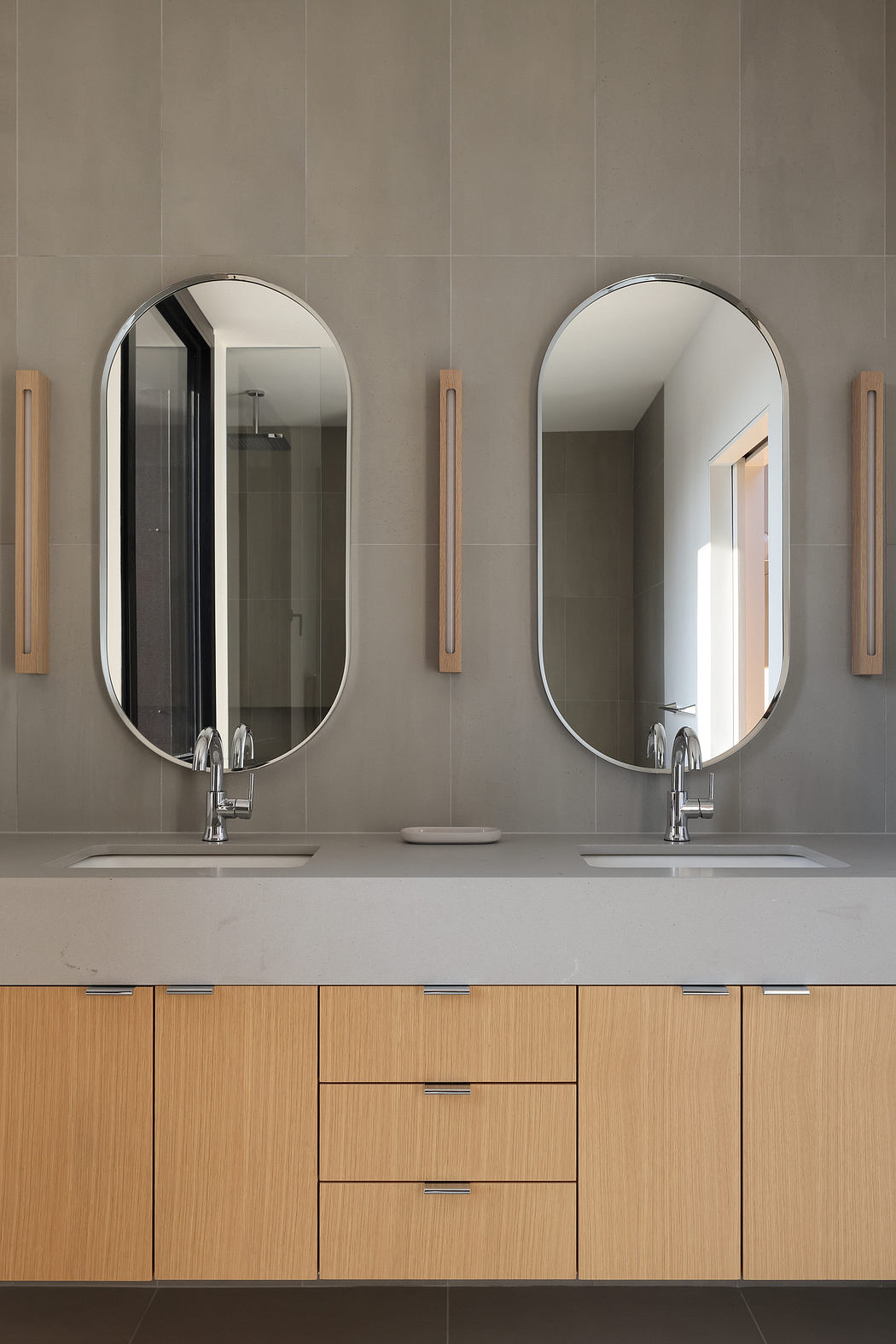 Minimalist bathroom vanity with oval mirrors, wood drawers, and wall-mounted light fixtures.
