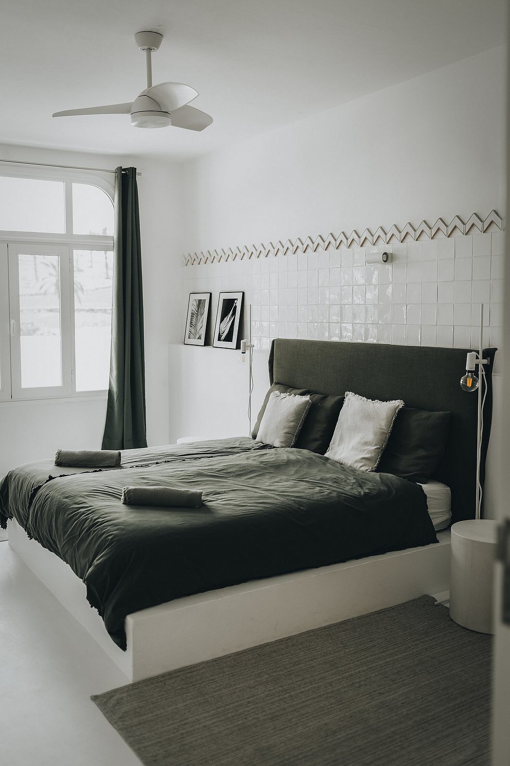 Minimalist bedroom with tiled walls, ceiling fan, and grey bedding on a white platform bed.