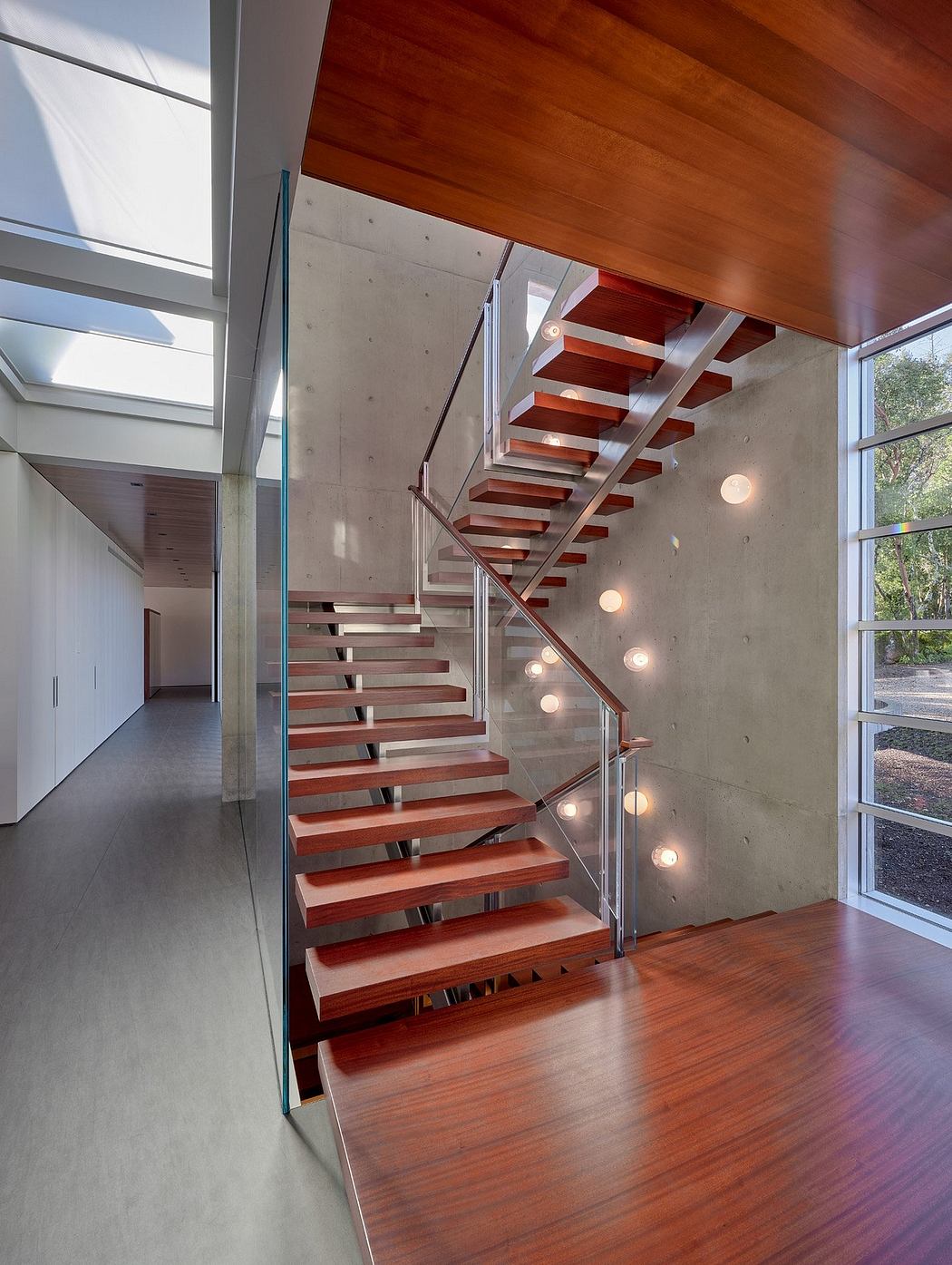 Modern staircase with wooden treads, concrete walls, and glass railings in a bright, airy interior.