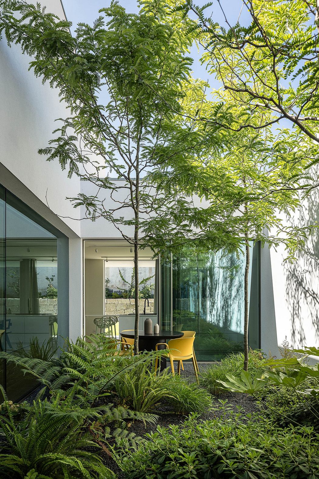 A lush, verdant garden surrounds a modern, glass-enclosed living space with yellow chairs.