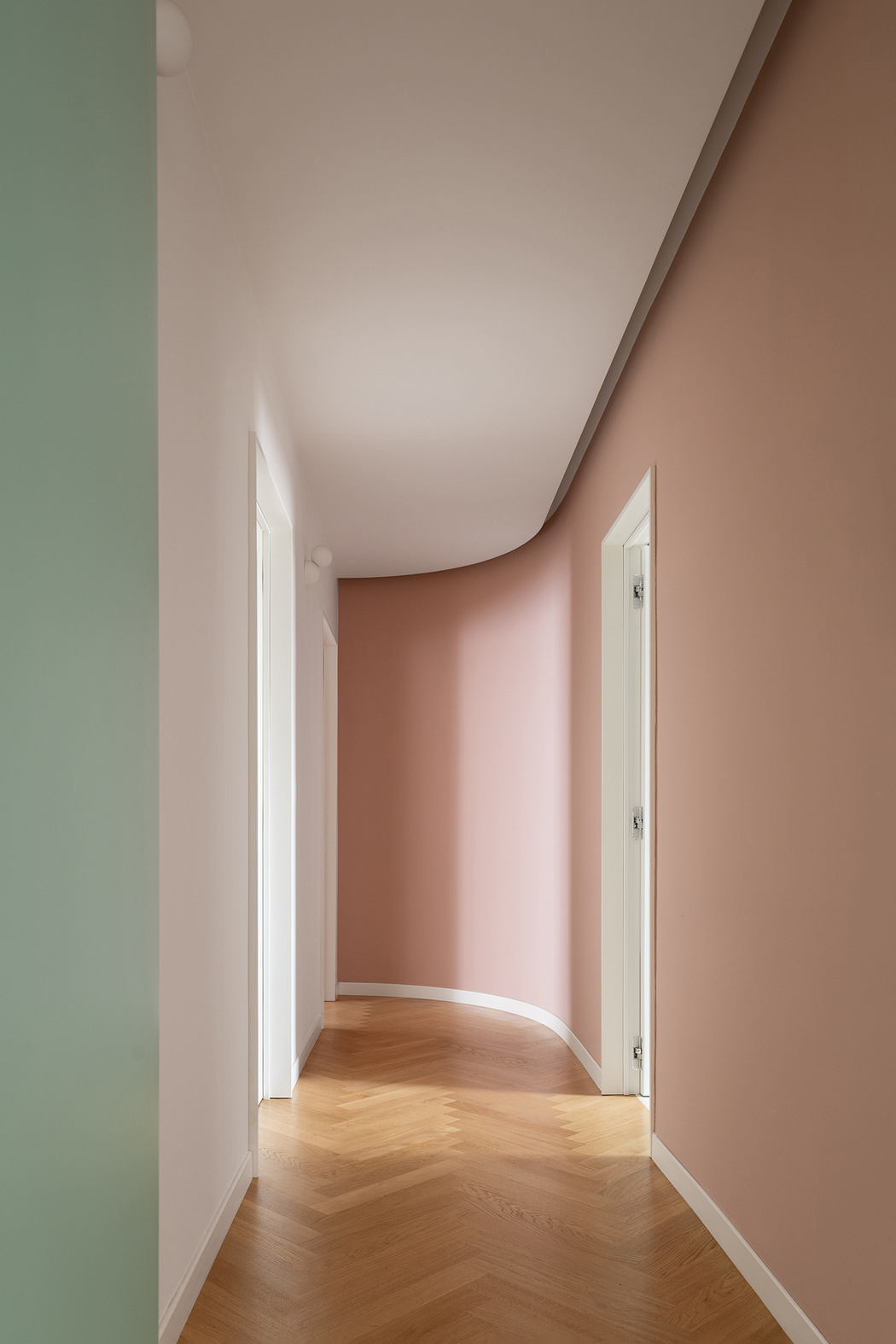 A minimalist hallway with curved walls in soft pink hues and a herringbone wood floor.