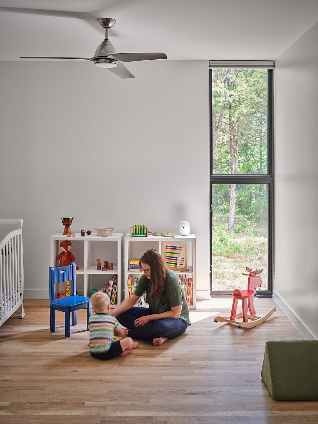 A bright, modern nursery with built-in shelves, a ceiling fan, and a large window overlooking lush greenery.