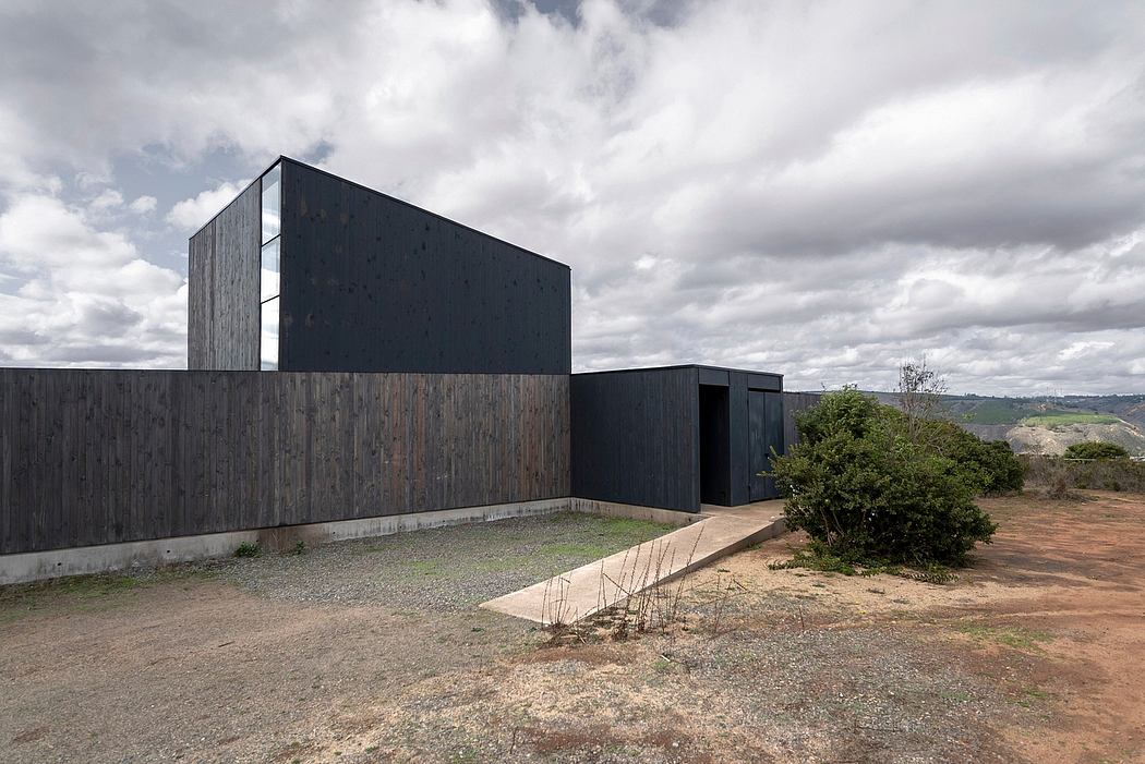 A modern black building with wooden panels, surrounded by a gravel path and vegetation.
