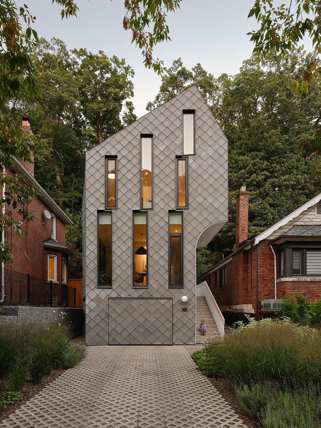 A modern, angular home with a unique tiled facade and prominent windows, surrounded by lush vegetation.
